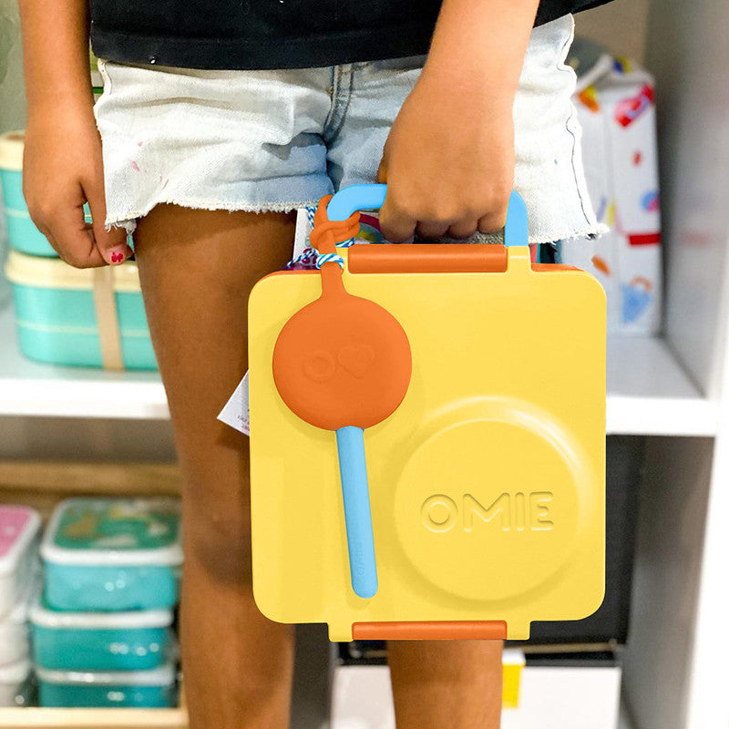 Yellow lunch box with orange handle and blue spoon held by a person in a store setting