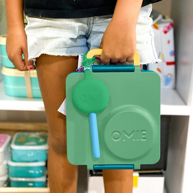 Person holding a green OMIE lunch box with a straw in a store setting
