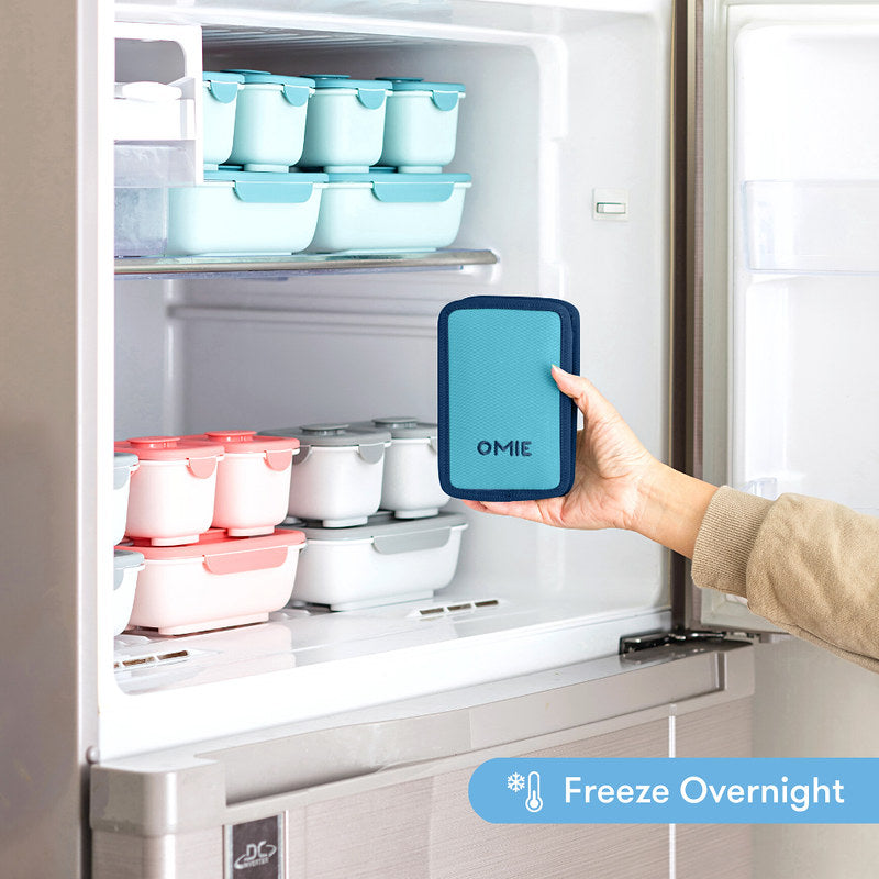 Hand holding a blue OMIE product in front of a refrigerator with food containers.
