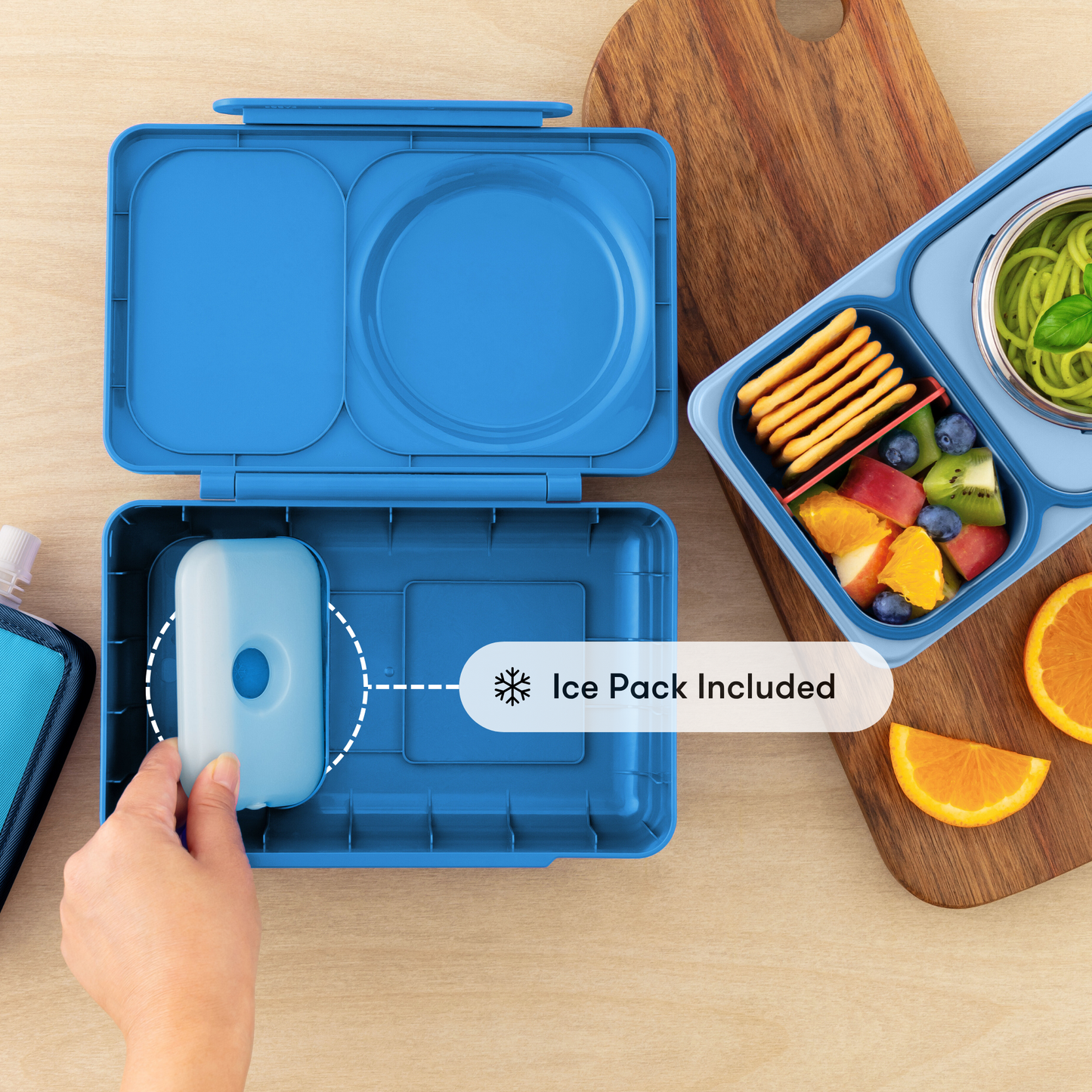 Blue bento box with ice pack included, next to a lunch setup with fruits and cookies on a wooden surface.