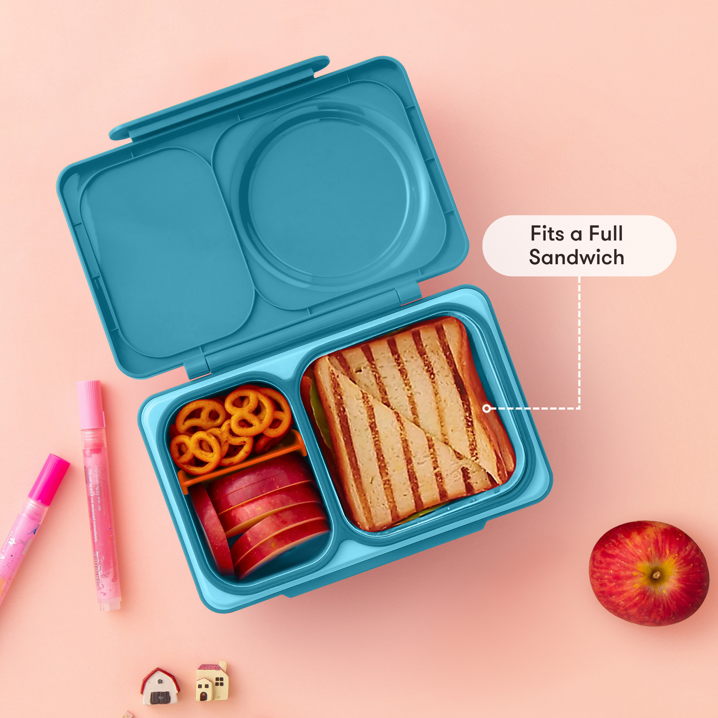 Blue bento box with compartments containing a sandwich, apples, and pretzels on a pink background.