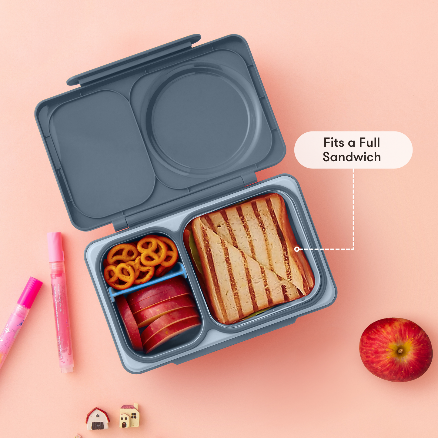 Gray bento box with compartments containing a sandwich, apples, and pretzels on a pink background.