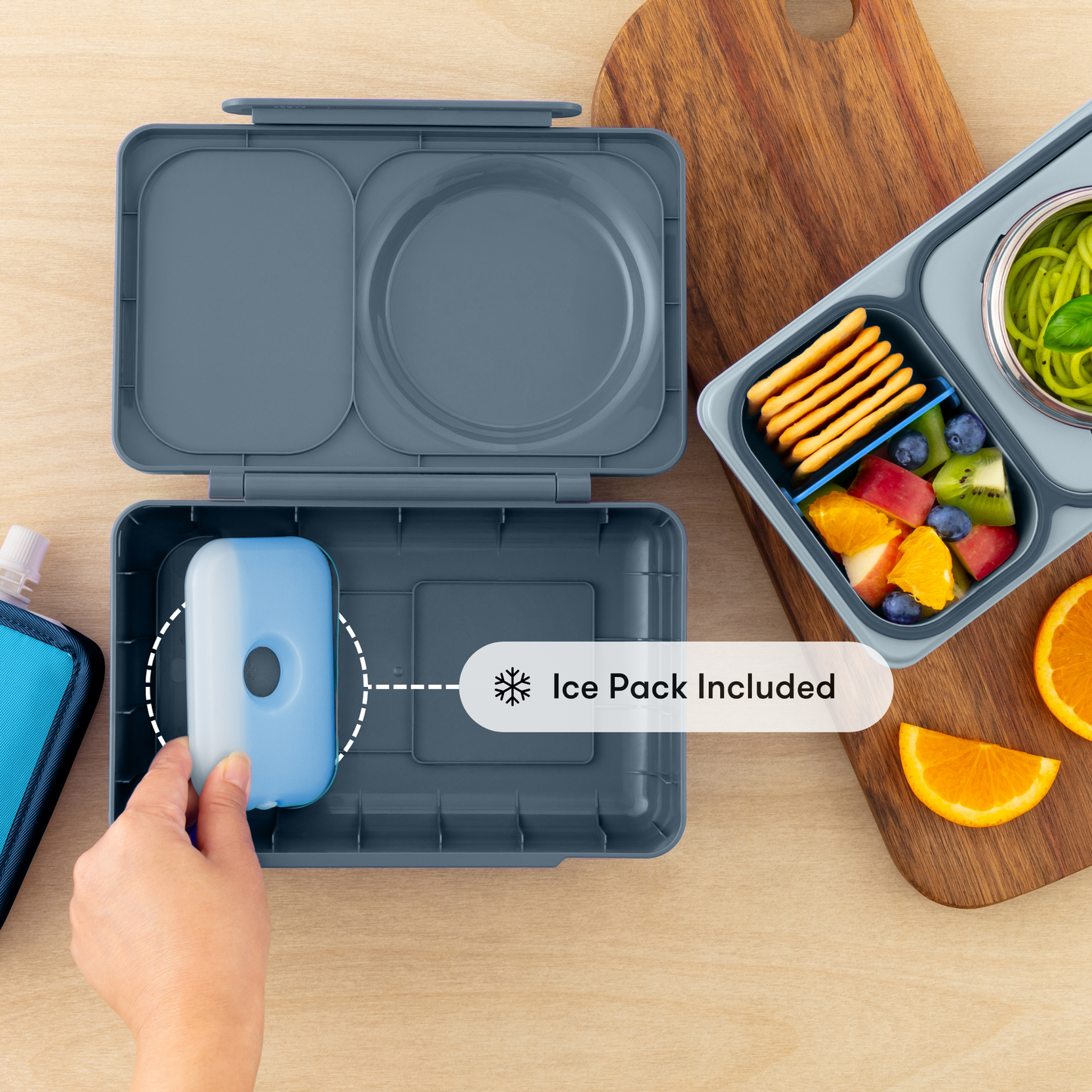 Gray bento box with ice pack included, surrounded by snacks and fruits on a wooden surface.