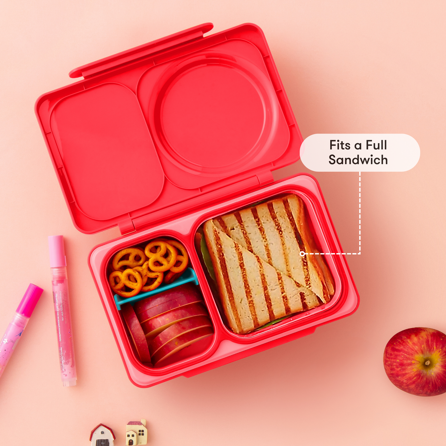 Red bento box with compartments containing a sandwich, apple slices, and noodles on a pink background.