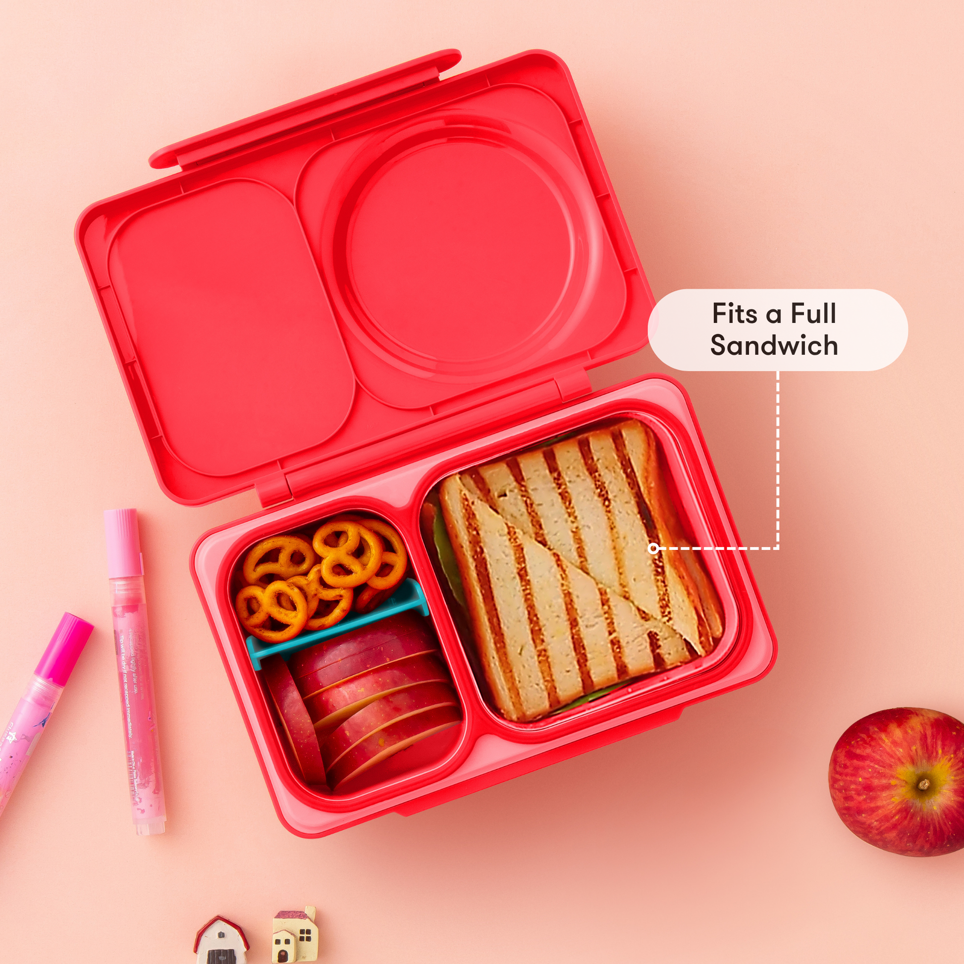 Red bento box with compartments containing a sandwich, apple slices, and noodles on a pink background.